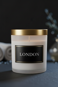 A frosted glass candle jar with a gold metal lid and a black label reading 'LONDON' in white text, set against a dark moody background with soft decorative